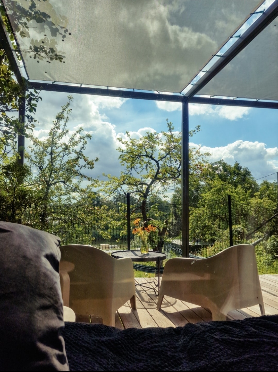 A shaded terrace is visible, featuring two white chairs and a small table. Sunlight filters through the canopy, illuminating the vibrant greenery outside. A vase with flowers adds a touch of color, while a view of fruit trees and a fenced garden is seen in the background.