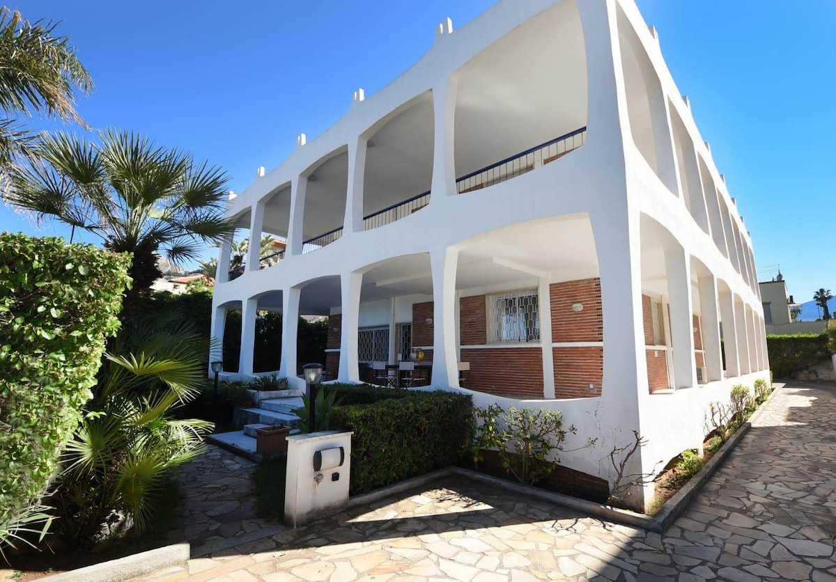 The exterior of the villa is showcased, featuring a white structure with multiple arches and a spacious terrace. Lush greenery surrounds the entrance, complemented by a stone pathway. Sunlight illuminates the scene, highlighting the architectural details and the inviting atmosphere of the property.