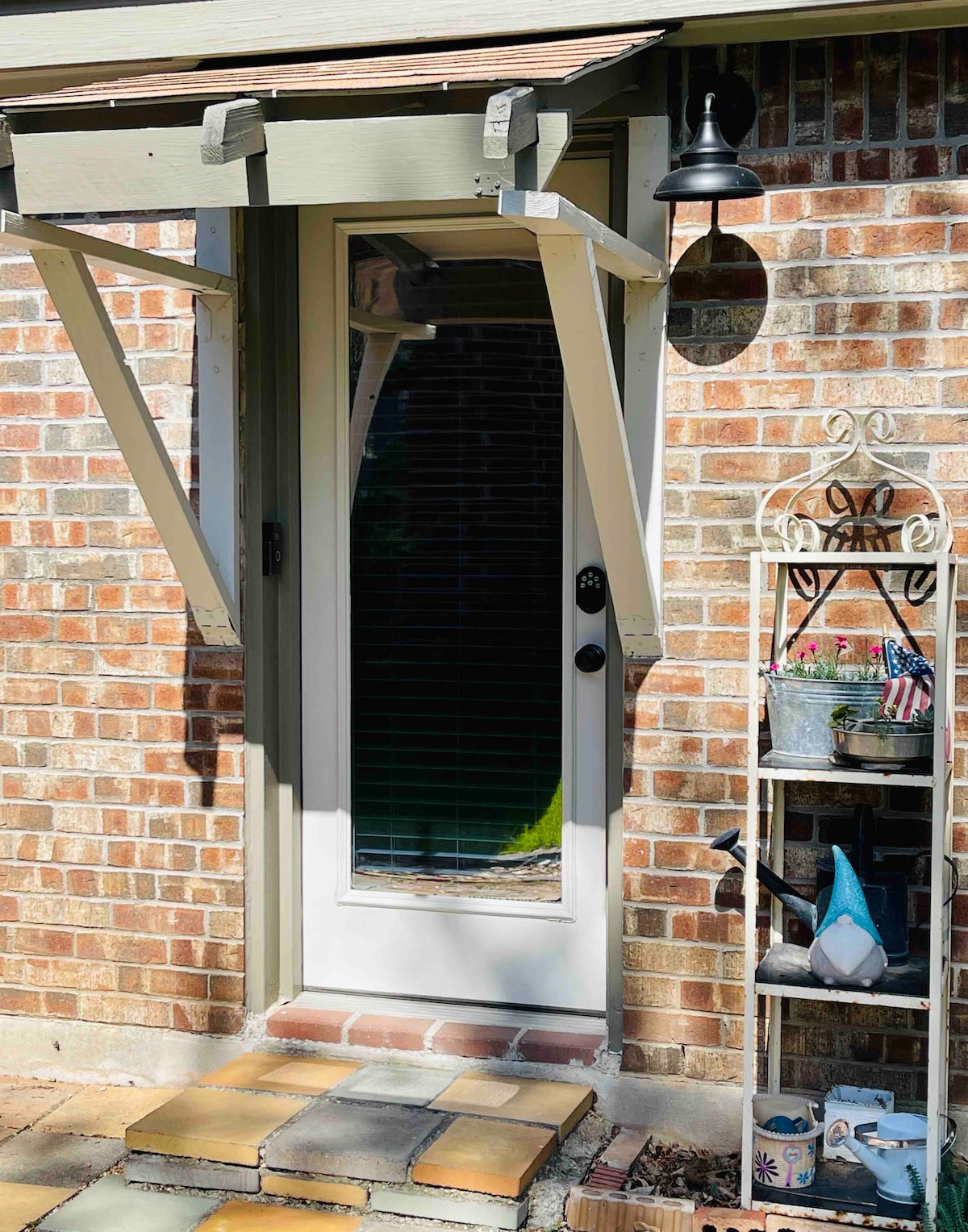 The image showcases a private entrance with a glass door framed by a light-colored overhang. Adjacent to the door, a decorative metal shelf holds potted plants and garden items, adding a welcoming touch. The brick facade provides a warm, textured background.