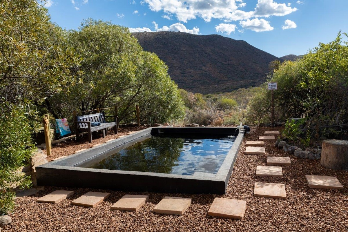 A natural splash pool is surrounded by stone pathways and landscaped with gravel. A bench is positioned nearby, offering a place to relax. Lush greenery frames the area, with distant mountains visible under a sky dotted with clouds.