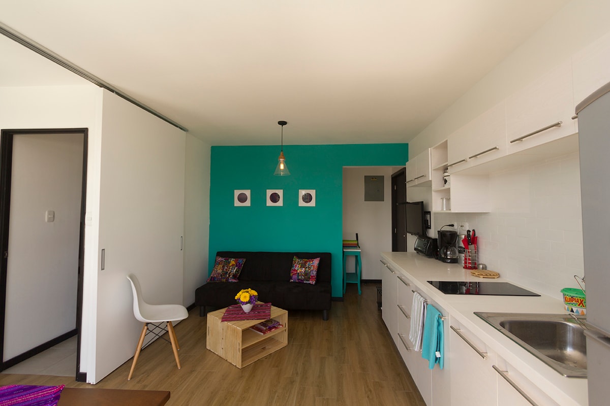 A bright apartment interior features a modern kitchen with white cabinetry and a stainless steel sink. A dark sofa, adorned with colorful cushions, complements the turquoise accent wall. Natural light fills the space, highlighting the open layout and wooden flooring.