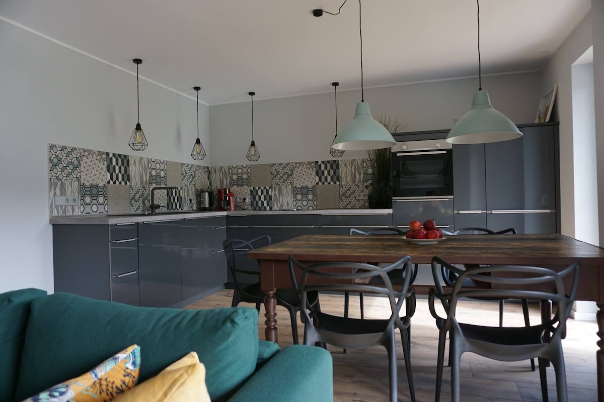 A modern kitchen features sleek dark cabinetry and stylish pendant lighting. A spacious dining table with eight chairs is positioned nearby, complemented by decorative elements. The backsplash showcases intricate patterned tiles, adding a unique touch to the room's overall design.