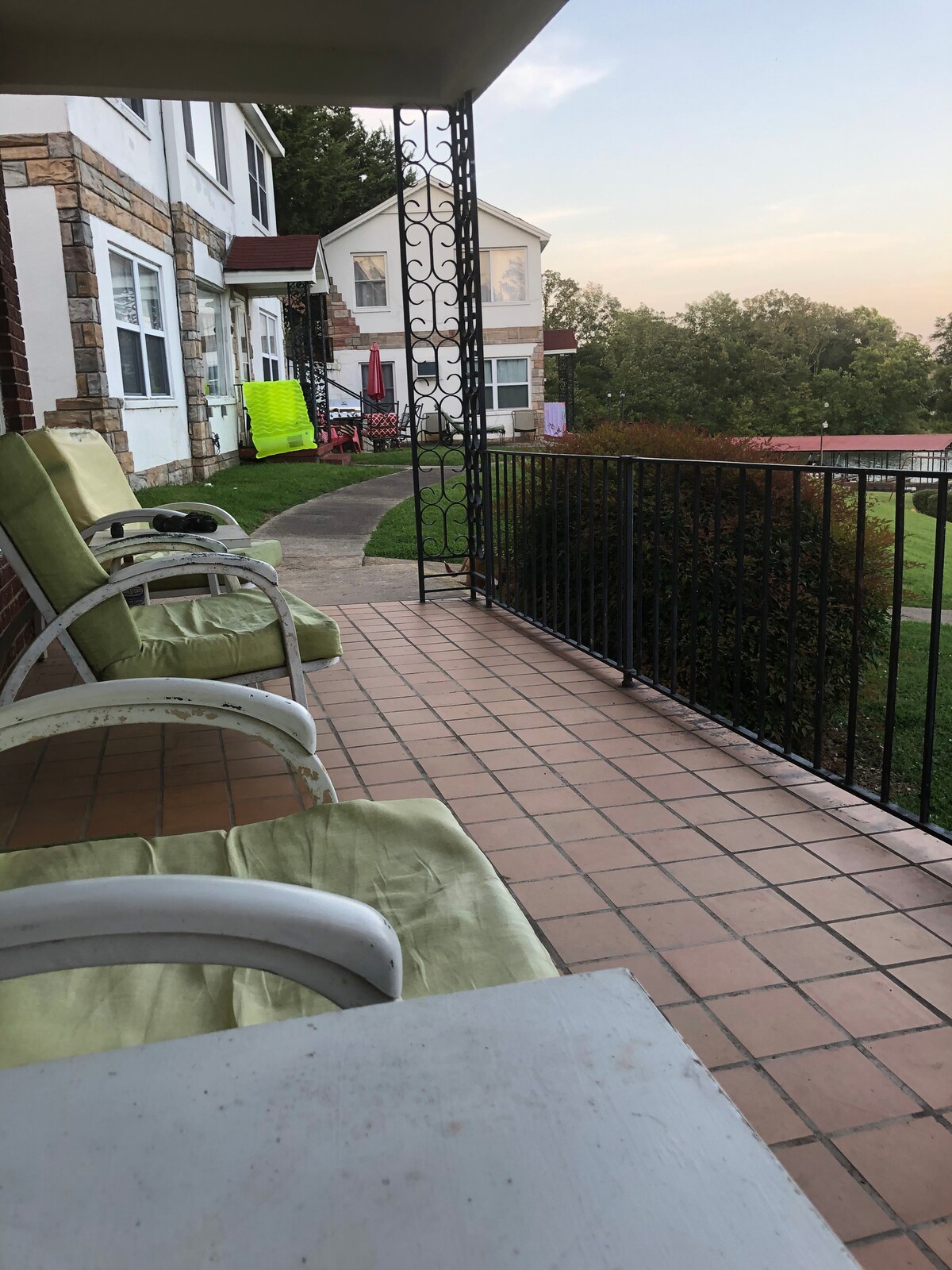 A porch area is seen featuring several light green chairs arranged around a small table. A view of the lush lawn is visible beyond the railing, with a pathway leading towards the lake. The setting captures a calm atmosphere during early evening light.