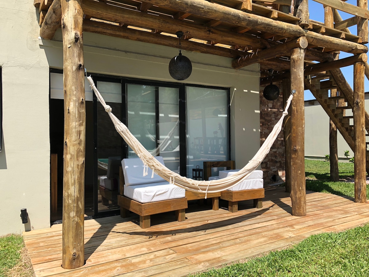 A wooden deck features a comfortable hammock suspended between sturdy posts. Two simple wooden lounge beds provide additional seating. Large glass doors lead into the interior space, bringing in natural light and a view of lush green surroundings.