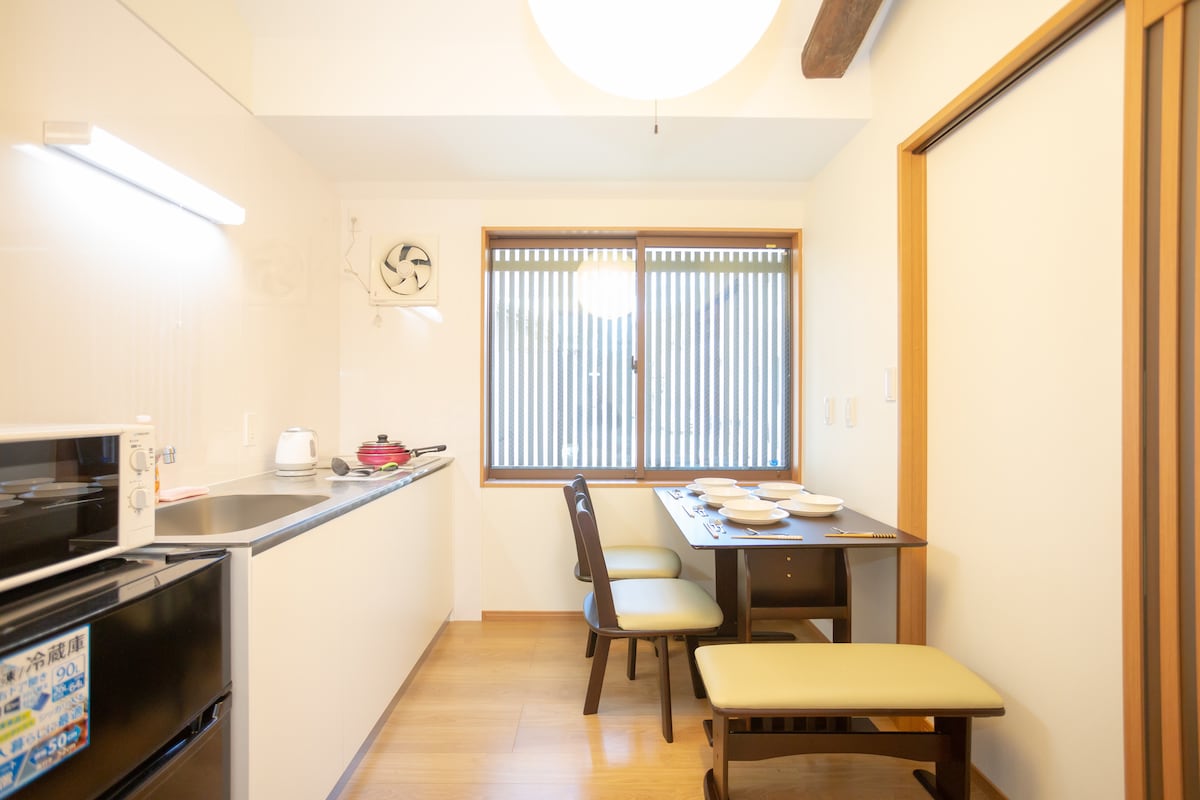 A compact kitchen area is equipped with modern appliances, including a microwave and a stovetop. A dining table with seating for four is situated near a window adorned with horizontal blinds, allowing natural light to fill the space.