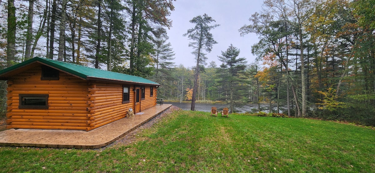A log cabin features a rustic exterior with a green roof, nestled among trees. A spacious lawn extends toward a serene body of water, while two wooden chairs are positioned on the patio, offering a peaceful view of the natural surroundings.