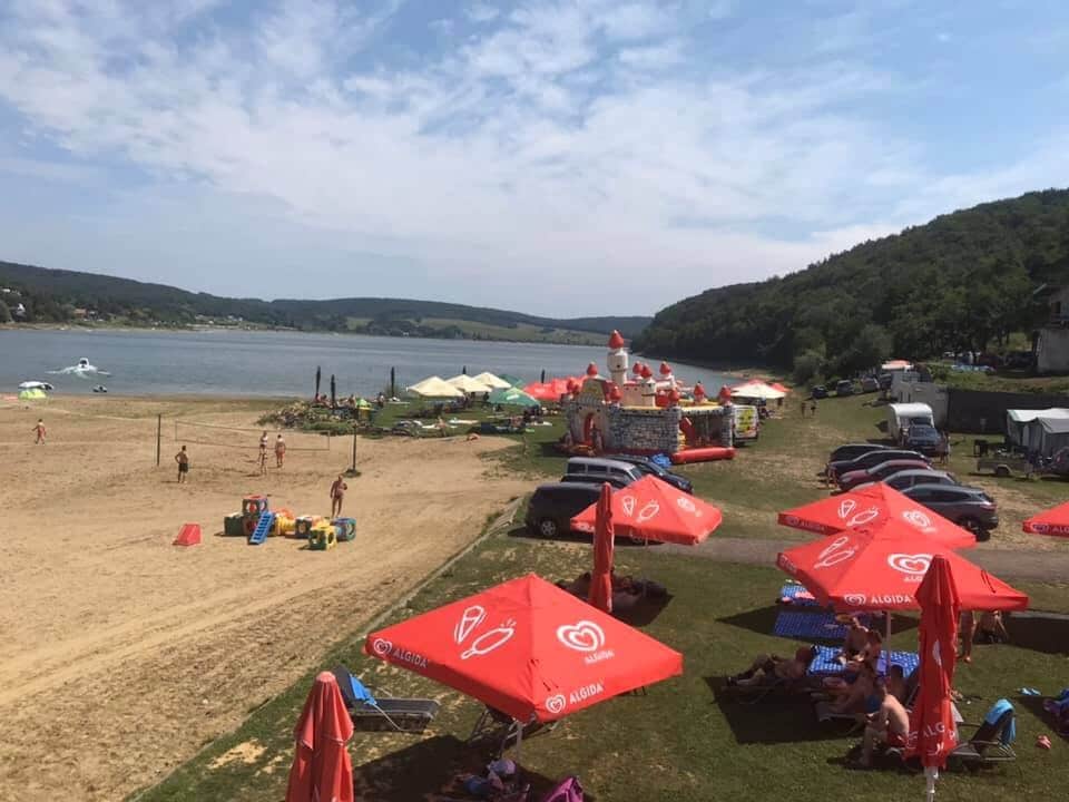 A sandy beach is visible with sun umbrellas providing shade to guests. Colorful tents line the shoreline, while people can be seen enjoying the water and recreational activities. The serene lake and surrounding hills create a calm backdrop to the beach area.