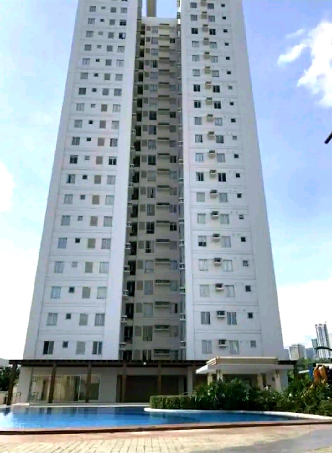 A tall residential building is presented, featuring a sleek façade with multiple windows. The entrance area includes a shaded outdoor seating space, and a swimming pool is visible in the foreground, surrounded by green landscaping, contributing to a modern urban atmosphere.