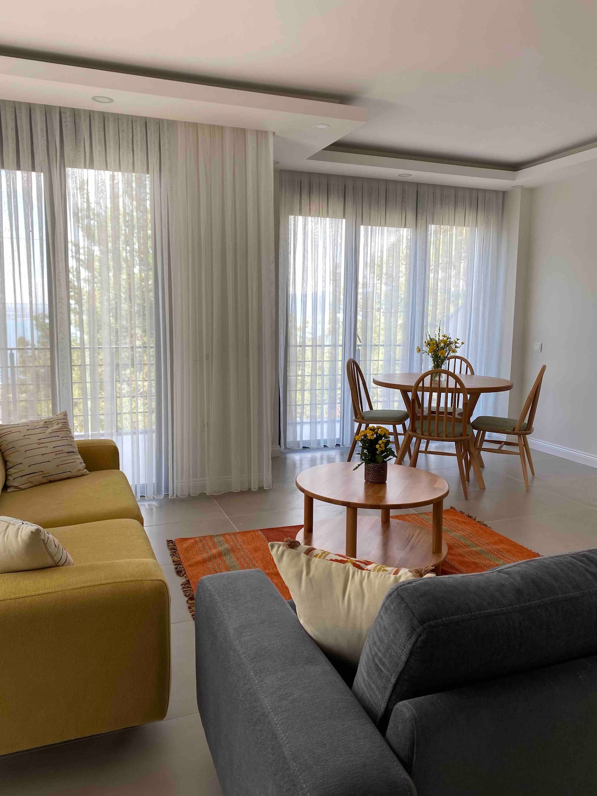 A modern living area features two comfortable sofas positioned around a circular coffee table. Natural light filters through floor-to-ceiling sheer curtains, creating a bright atmosphere. In the background, a dining table with wooden chairs is set against a view of greenery through large windows.