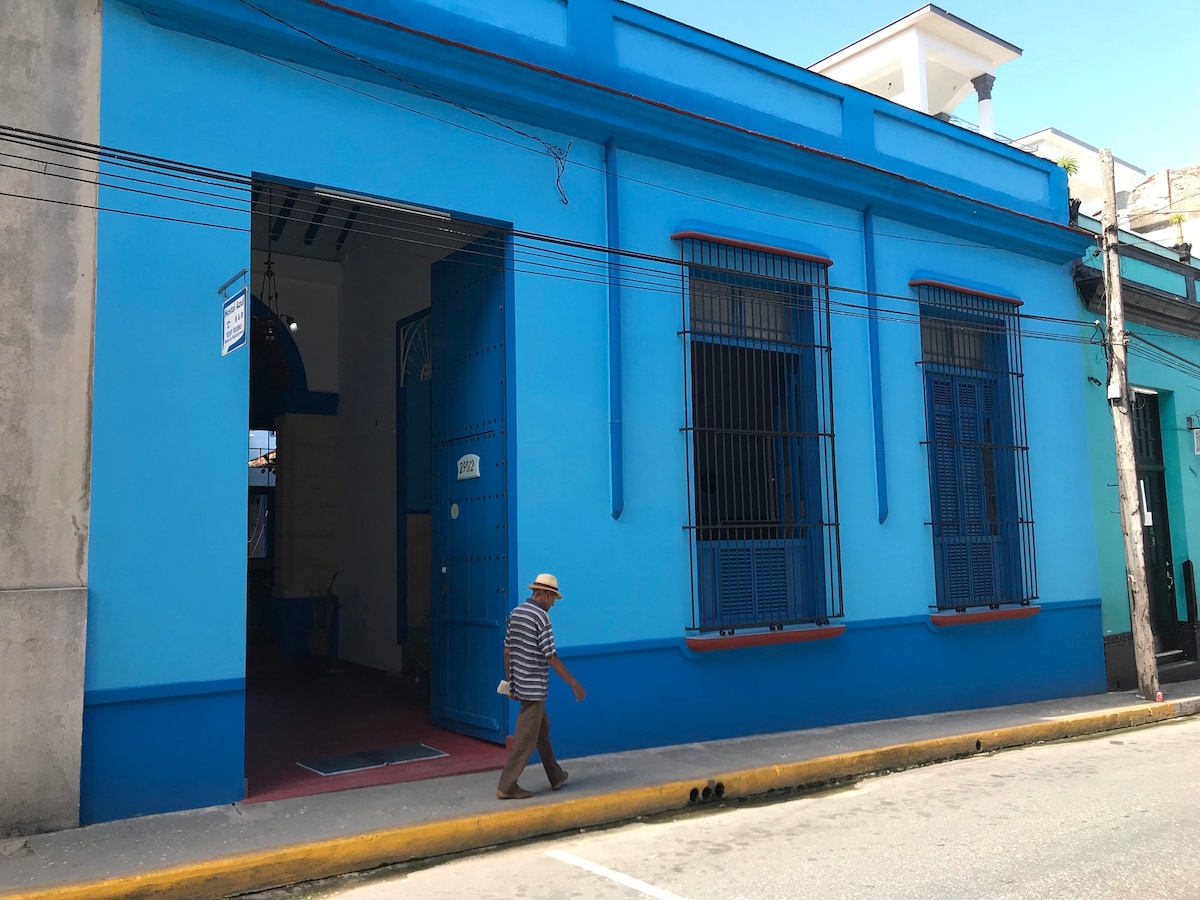 The exterior of a vibrant blue colonial house is shown, featuring large, arched doors and tall windows with metal grilles. A pedestrian walks past the building, highlighting the lively atmosphere of the street in front. Details of the bright façade are accentuated by natural light.