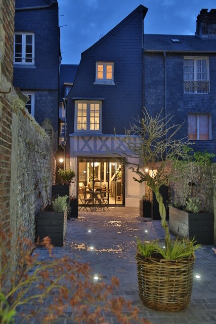 An evening view captures the façade of a charming house, with its dark roof and distinctive architectural lines. Soft lighting illuminates the paved courtyard, where planters with greenery are placed. Large glass doors invite entry, revealing the interior space beyond.