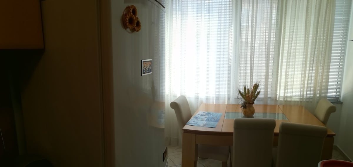 A dining area is featured with a wooden table at its center, surrounded by four light-colored chairs. Natural light filters through sheer white curtains, illuminating a floral centerpiece on the table. A refrigerator stands nearby, completing the kitchen space.