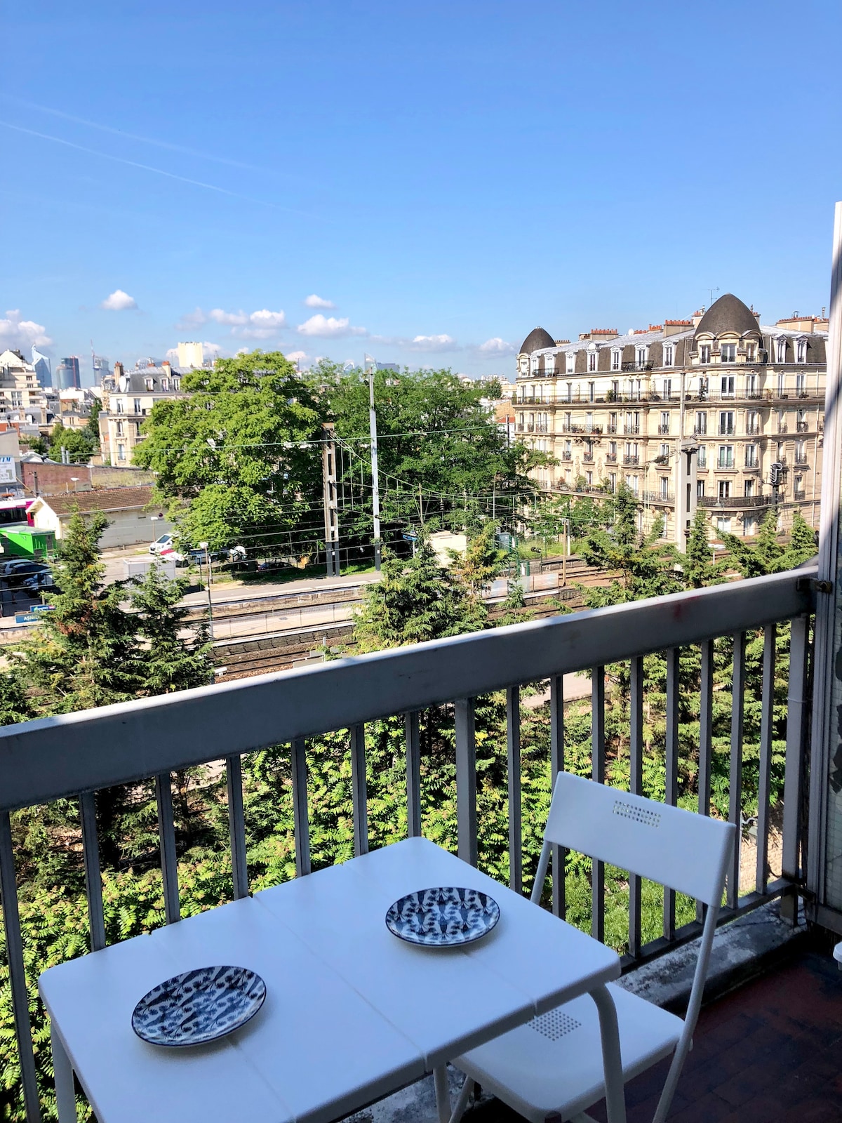 A balcony is shown with a small white table and two chairs, set against a backdrop of greenery and city buildings. The views include trees and distant architecture, under a clear blue sky with scattered clouds.