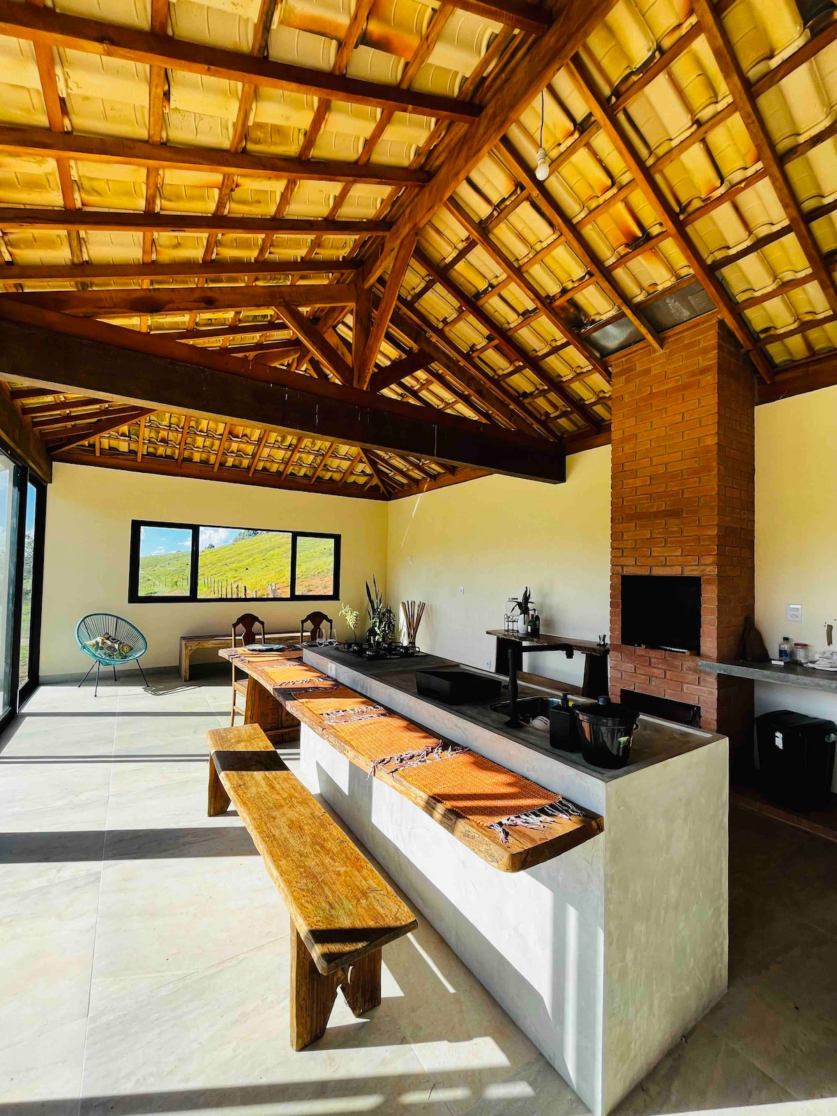 An open kitchen area features a high, wooden ceiling with exposed beams. A rustic wooden bench runs alongside a stone countertop, equipped with a grill. Large windows provide views of the surrounding greenery, enhancing the spacious atmosphere.