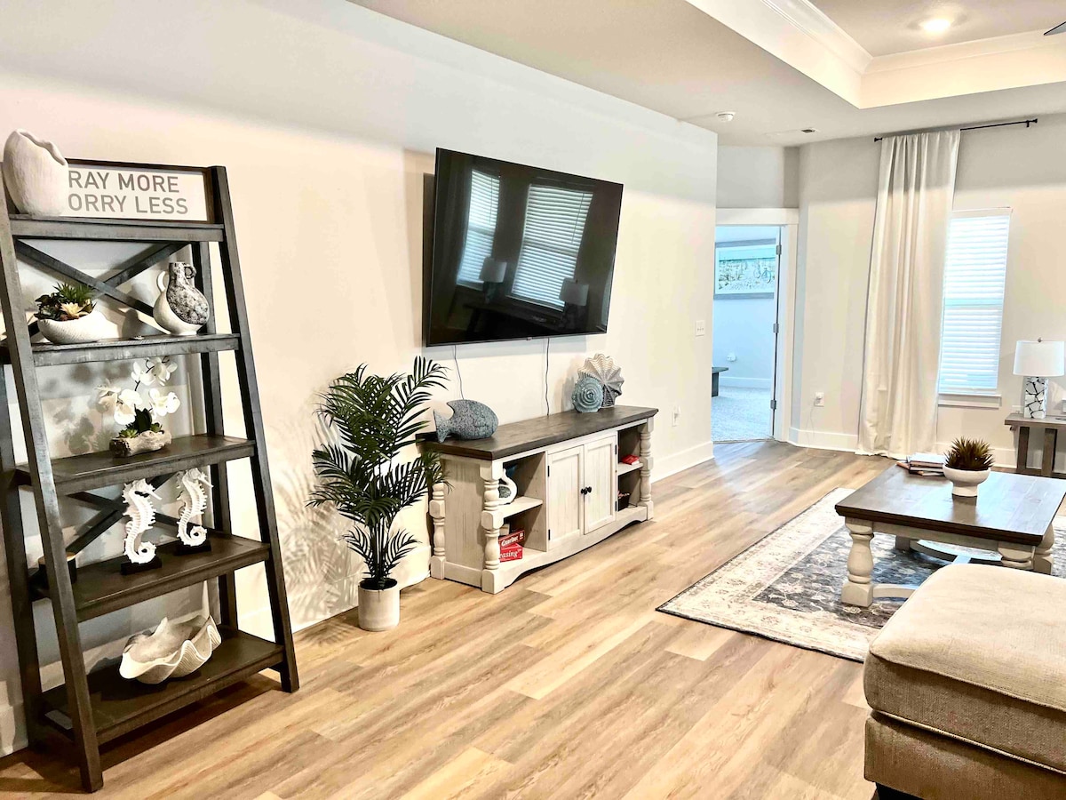 A cozy living area features a spacious sectional sofa and a mounted television. A wooden shelf displays decorative items, while a potted plant adds a touch of greenery. Natural light filters through sheer curtains, creating a bright and welcoming space.