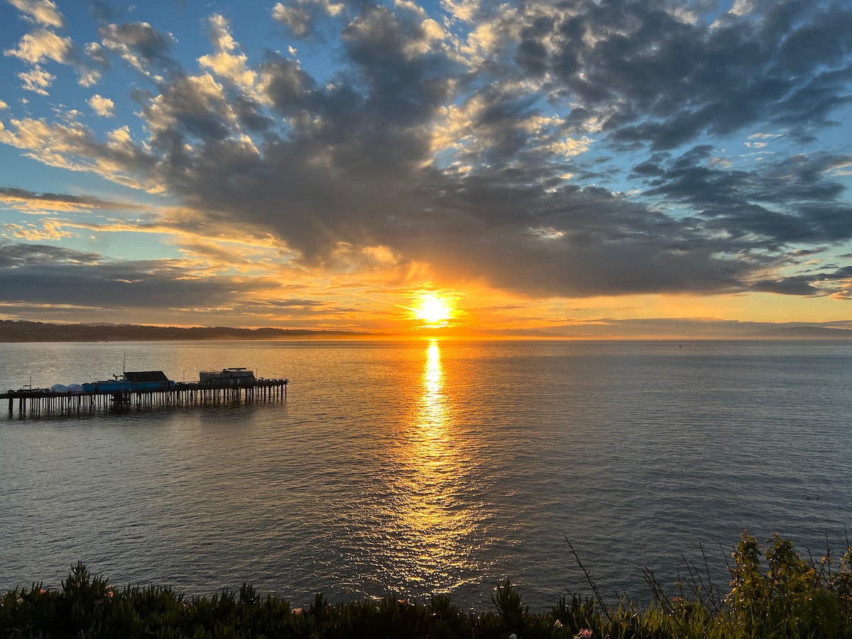 A serene view of the ocean during sunset features warm golden hues reflecting on calm water. A pier extends into the sea, framed by soft clouds in the sky. The tranquil scene suggests a peaceful coastal atmosphere.