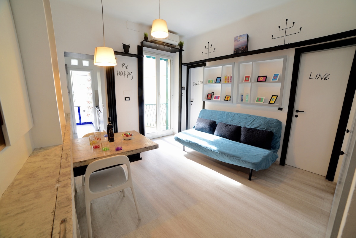 A bright and airy living area features a blue sofa bed and a wooden dining table with white chairs. Wall shelves display colorful photos, and two pendant lights hang from the ceiling. Large windows allow natural light to fill the space, enhancing the room's welcoming feel.