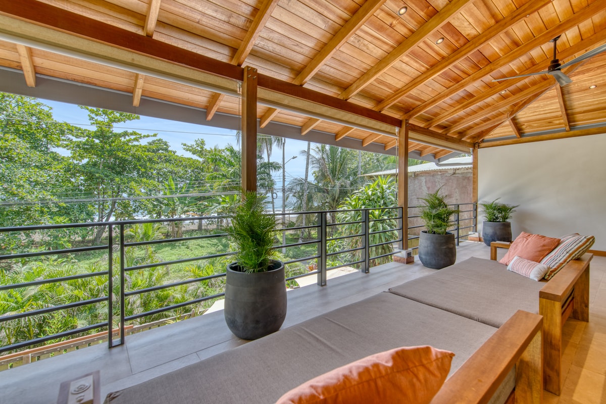 A spacious terrace features comfortable lounge seating and two potted plants. Wooden beams and a high ceiling enhance the area, while large sliding glass doors offer views of lush greenery and the nearby beach. Natural light fills the space, creating an open and airy ambiance.