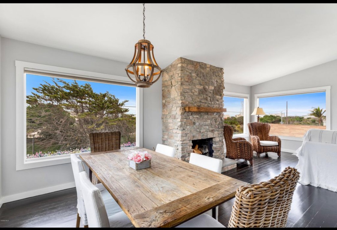 A dining area is highlighted by a wooden table surrounded by six chairs, with two wicker armchairs positioned beside a stone fireplace. Expansive windows frame the room, providing views of the natural landscape and enhancing the inviting atmosphere.