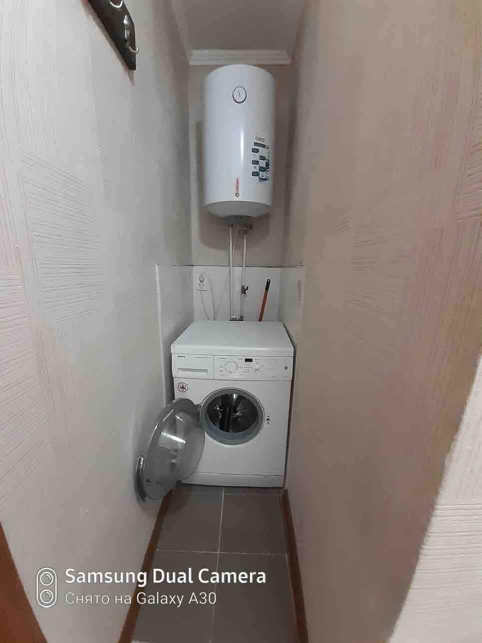 A compact laundry area is visible, featuring a washing machine and a wall-mounted water heater. The space is surrounded by light-colored walls and tiled flooring, providing a functional environment for laundry needs.