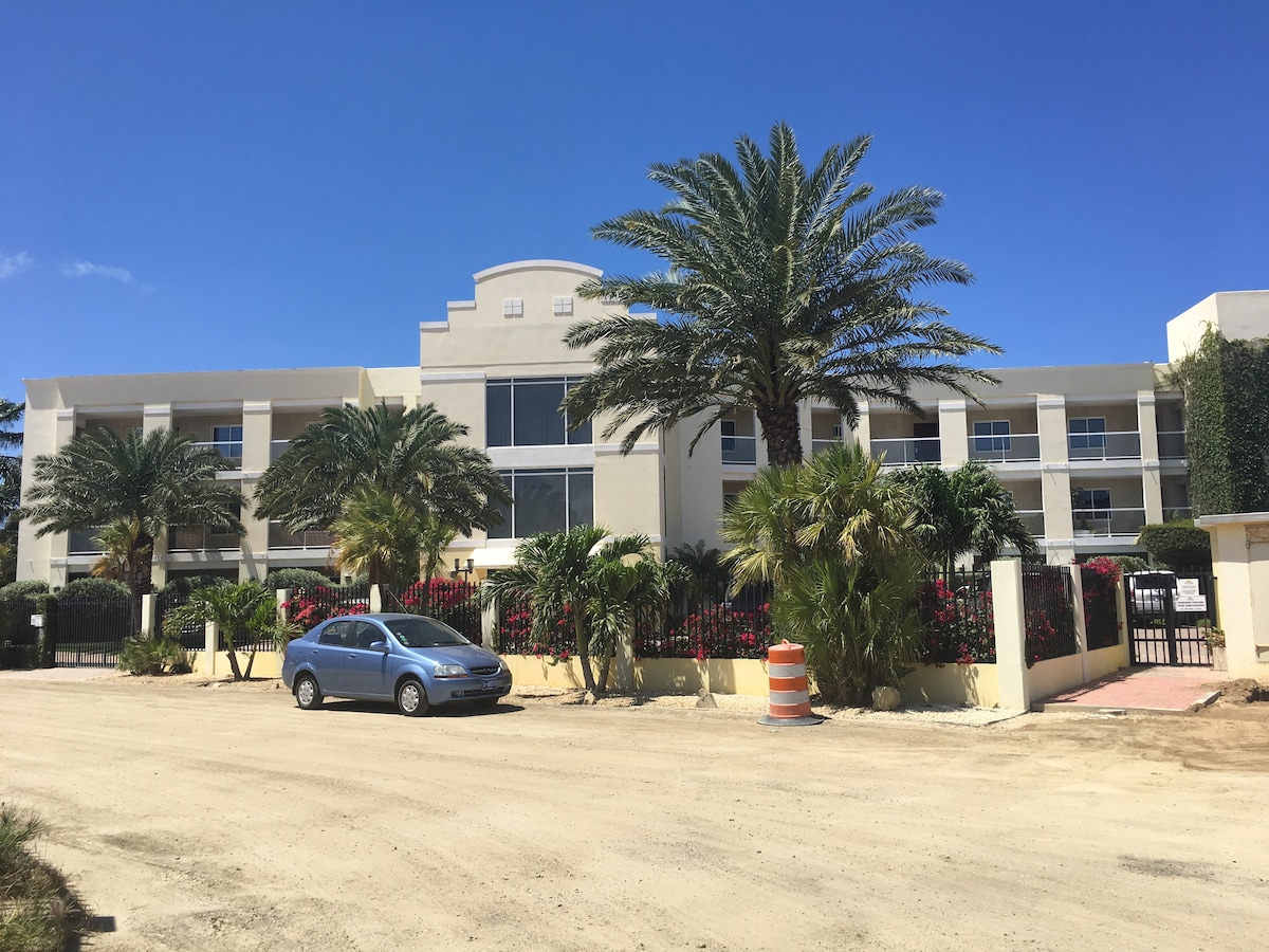The exterior of a modern three-story building is displayed, framed by palm trees and tropical plants. A gravel driveway leads to the entrance, with a parked car nearby. Bright blue skies serve as a backdrop, enhancing the inviting appearance of the residence.