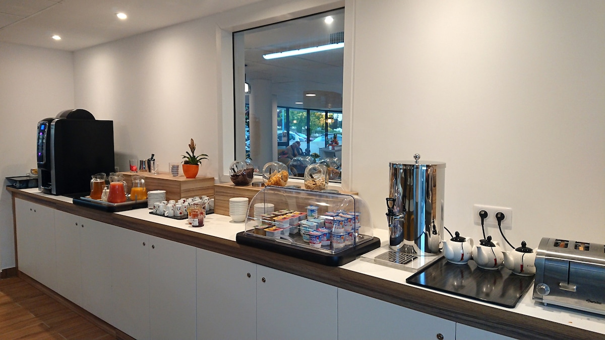 A modern kitchenette is presented with a variety of food items on display. Shelves hold jars of preserves, cereal containers, and a coffee machine. A glass case showcases pastries and assorted snacks. A large window allows natural light to illuminate the space.