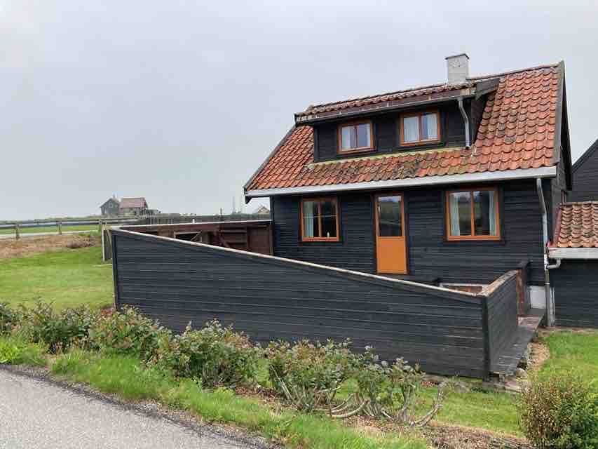 A traditional two-story cabin is showcased, featuring dark wooden siding and a red-tiled roof. The front door is visible, framed by large windows that allow ample natural light. Surrounding greenery is seen, creating a serene outdoor space.