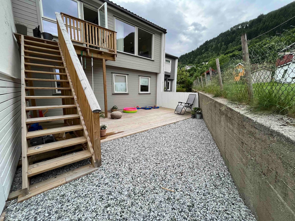 An outdoor space is presented with a wooden deck and a stone pathway. A set of stairs leads to the upper level, while lounging chairs are arranged nearby. Colorful mats and plants are visible, creating a relaxed environment. A grassy area and a fence outline the property.