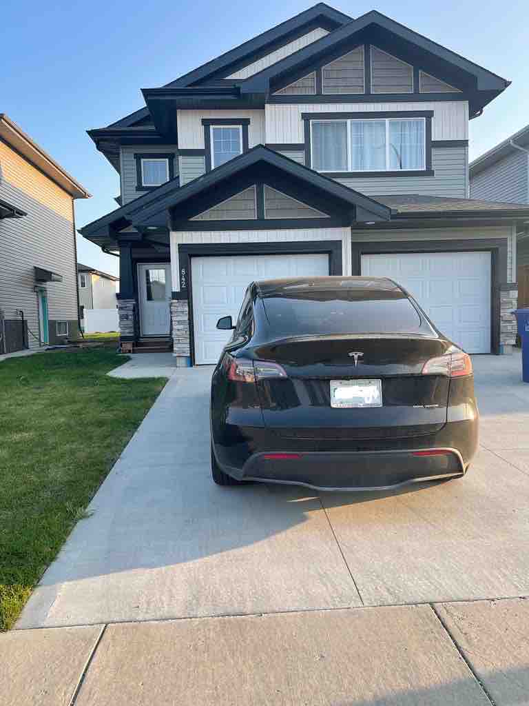 A modern two-story house features a front driveway accommodating a parked black electric vehicle. The exterior showcases a combination of light and dark siding with stone accents. A well-maintained lawn is visible, and the double garage doors are closed.