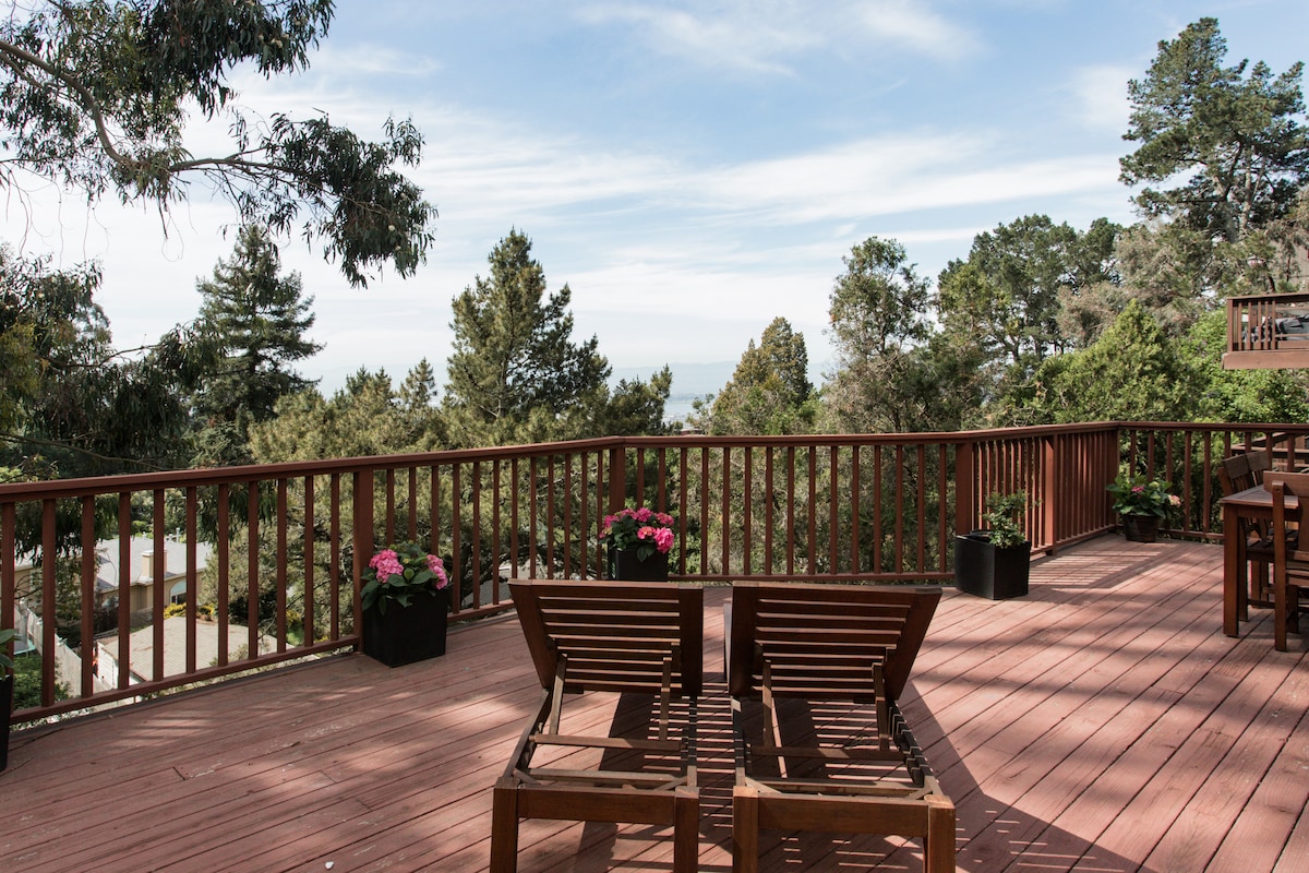 A spacious wooden deck features two lounge chairs positioned for enjoying views of the surrounding trees and distant water. Flower pots with blooming plants add a touch of color, complementing the natural setting.