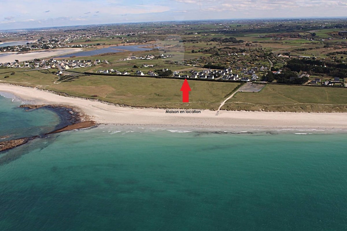 An aerial view showcases a tranquil coastline with soft sandy beaches meeting clear turquoise waters. In the background, a collection of houses is positioned near the shoreline, bordered by lush green fields and expansive open landscapes.