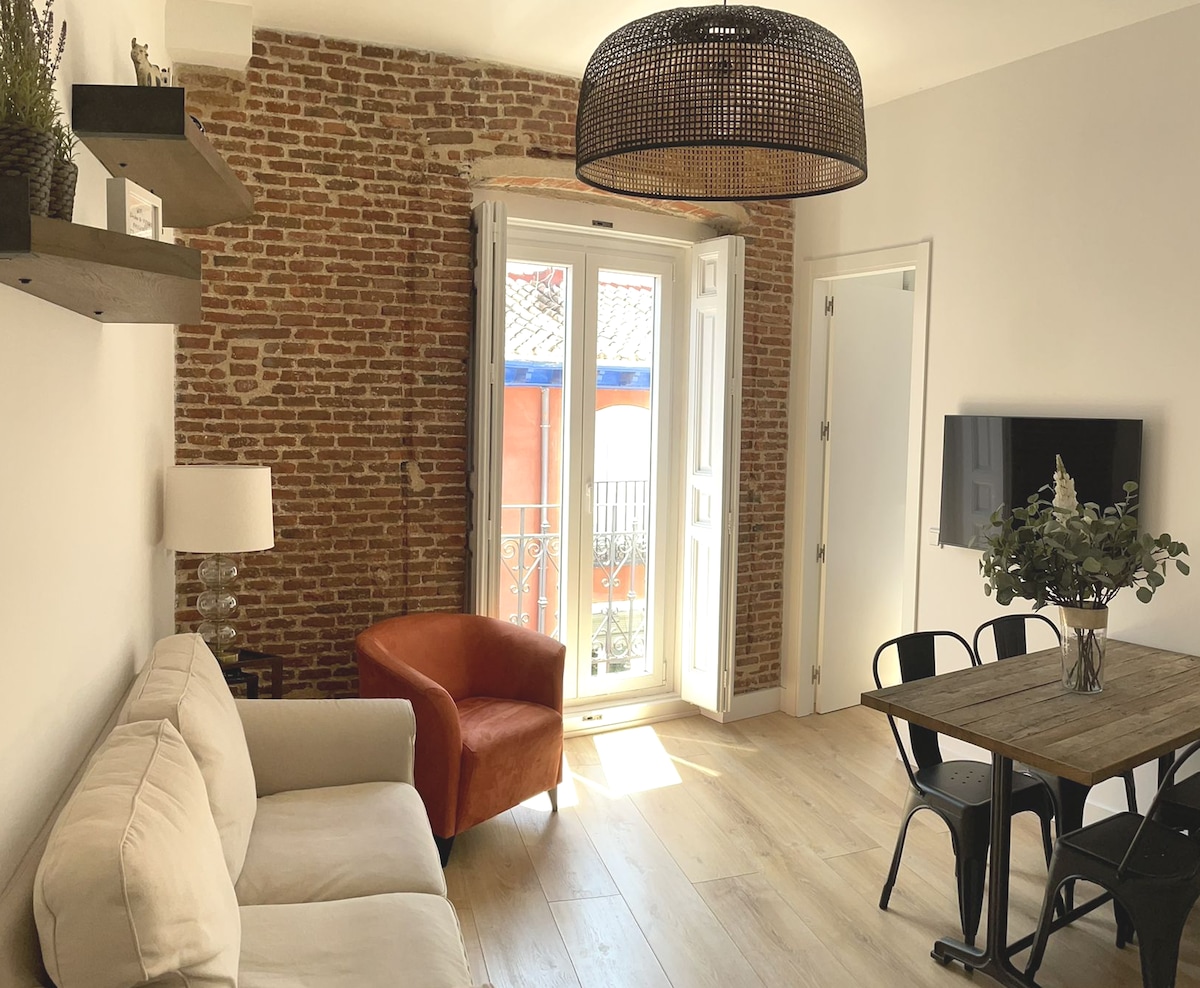 A living area features exposed brick walls and natural light streaming through a pair of balcony doors. A cream sofa and an orange armchair are positioned beside a wooden dining table with black chairs. A modern light fixture hangs from the ceiling.