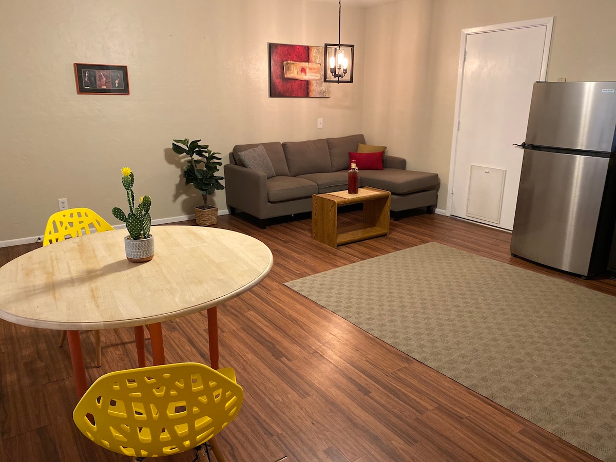 The living area presents a cozy configuration, including a spacious grey sofa facing a wooden coffee table. A round dining table with bright yellow chairs occupies one corner. A small potted cactus adds a touch of greenery, while the kitchen area is visible with stainless steel appliances.