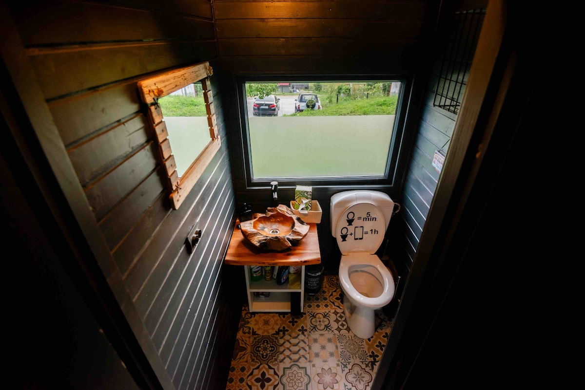 A compact bathroom features a toilet positioned beside a wooden countertop. A decorative mirror rests on the wall, and a window offers a view of the greenery outside. Unique patterned tiles cover the floor, adding character to the space.