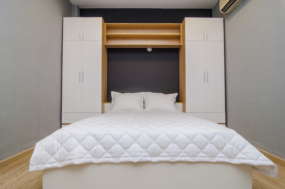 A spacious bedroom features a neatly made bed with a white quilt. Simple white cabinetry is located above the bed, providing ample storage. A dark accent wall adds a touch of contrast, while a light wooden floor completes the room's modern aesthetic.