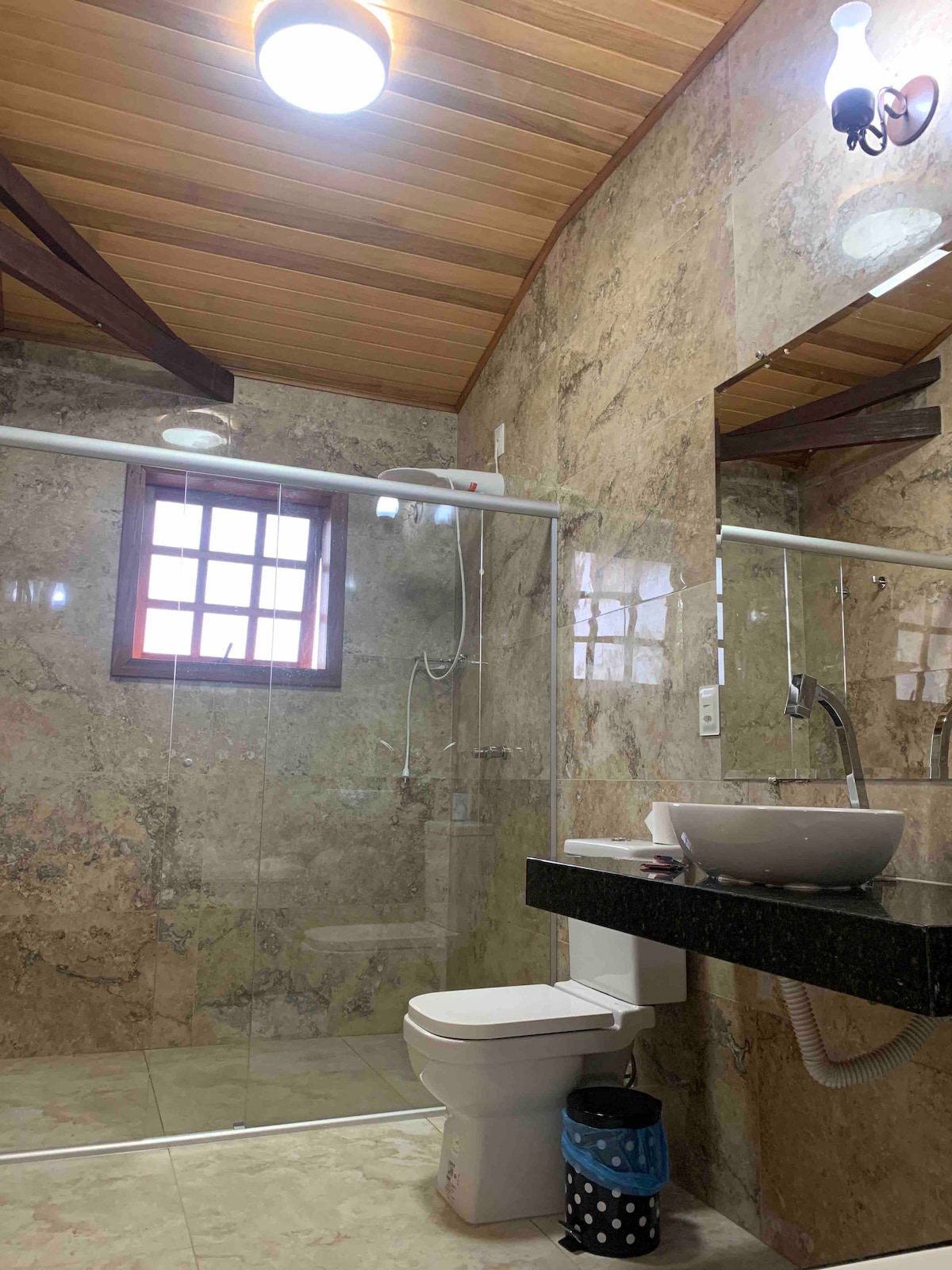 A modern bathroom is showcased with a glass shower enclosure and stone walls. A sleek black countertop with a white sink is visible, while a window allows natural light to enter. Additional fixtures, such as a mirror and toilet, are also present.