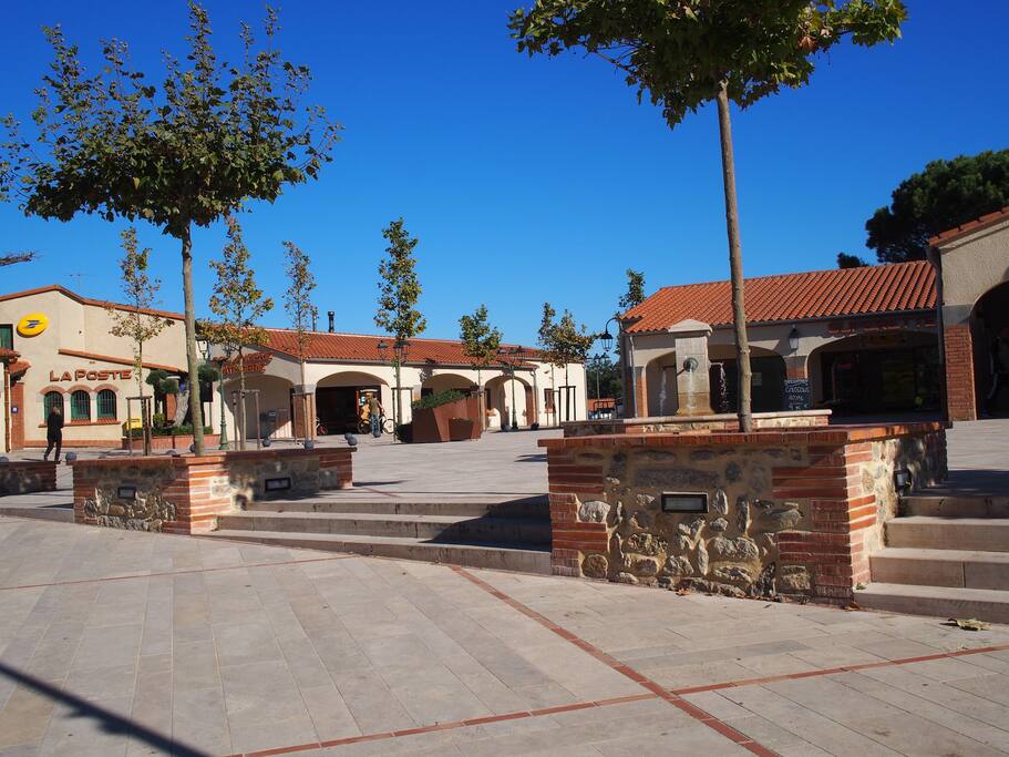 La place du village, avec mairie, poste, boulangerie, superette, presse-tabac et un grand parking derrière.
