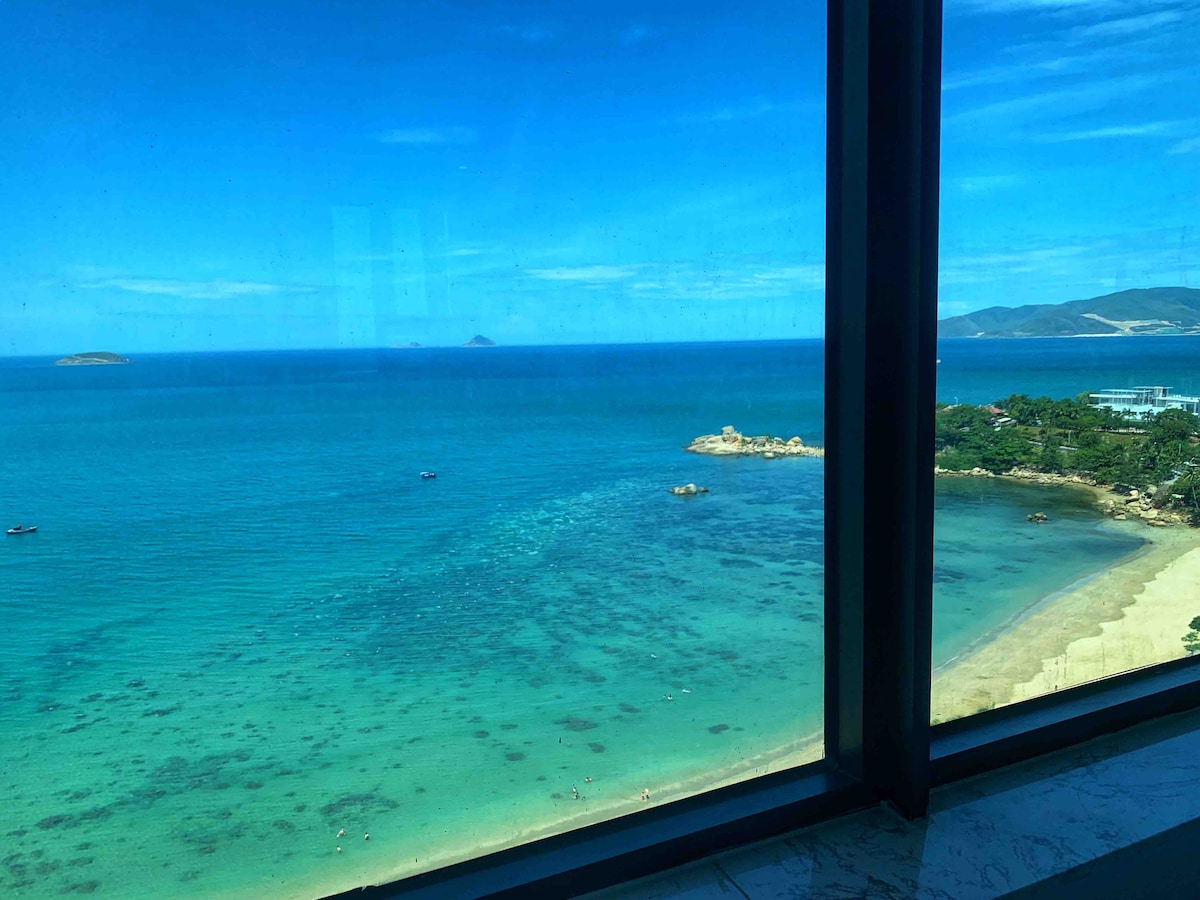 The image showcases a panoramic view of the beach and ocean from a large window. Clear turquoise waters stretch out to the horizon, with gentle waves lapping at the shore. A few boats are visible, enhancing the serene coastal landscape.