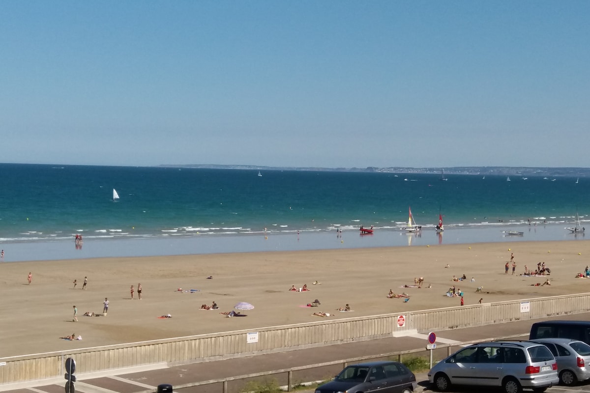 A wide beach stretches along the shore, with numerous beachgoers enjoying the sun. The calm blue sea is visible in the background, featuring small sailboats and a clear sky. The sandy area is marked with beach umbrellas and towels, creating a lively atmosphere.