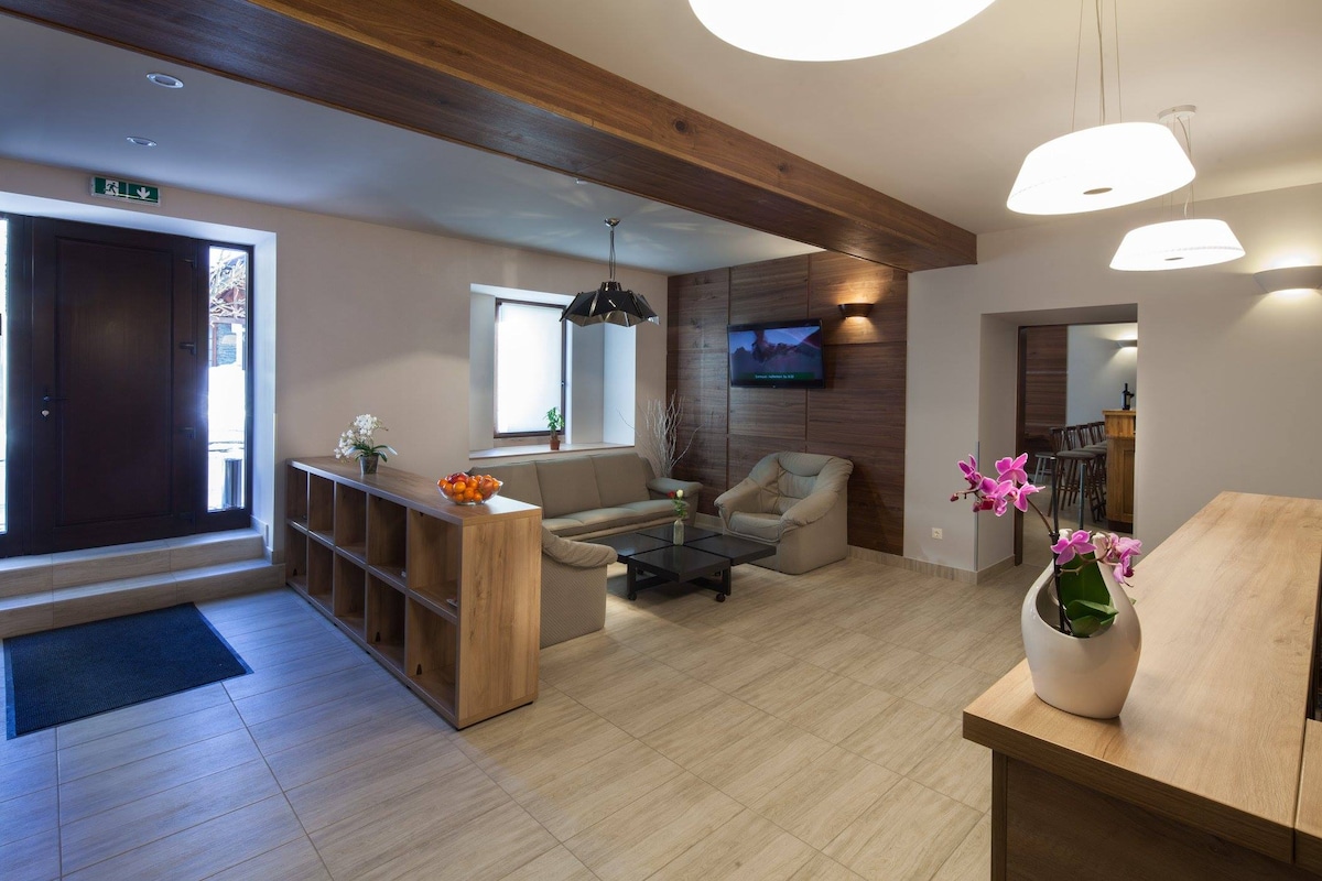 A welcoming lobby area features a modern design with wooden accents. A comfortable seating arrangement is provided near a coffee table, complemented by natural light from a nearby window. Decorative orchids and fruits add subtle touches to the atmosphere. A visible entrance leads outside, enhancing convenience.