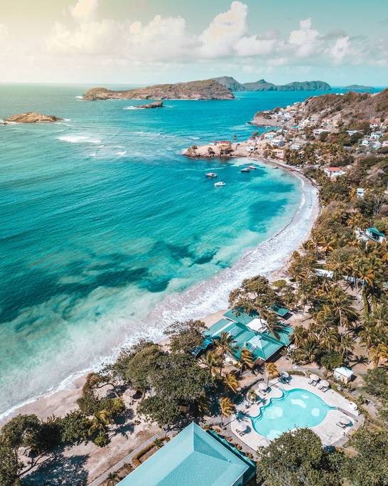 An aerial view captures a serene coastal landscape featuring a curved beach bordered by lush palm trees. The clear turquoise waters reflect the sky, while a pool with sun loungers is visible beside a shaded area. Distinct islands are seen in the distance.