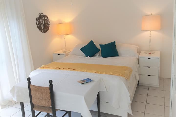 The room features a King Sized hand made wooden bed,  white cotton drapes and linens, bespoke bedside tables with charging strips, pretty lamps, Caribbean Art and a writers desk. Choice of ceiling fan or A/C (or both) to keep you cool. 