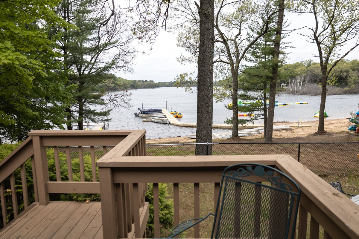A wooden deck offers a view of Kusel Lake, featuring a sandy beach and a dock with boats and kayaks anchored nearby. Lush trees frame the scene, providing a natural backdrop, while a metal chair is placed on the deck for relaxation.