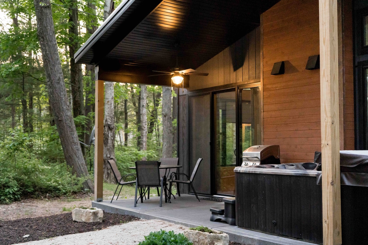 An outdoor patio area features a dining set with four chairs, alongside a grill and a hot tub. The area is surrounded by lush greenery, with tall trees providing natural shade. Soft evening light casts a warm glow over the space.