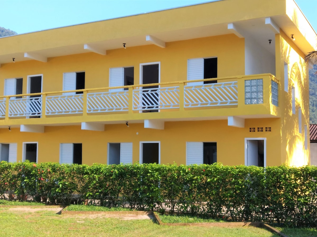 A bright yellow two-story building is showcased, featuring multiple balconies with white railings. The structure is surrounded by a tidy green hedge, and the clear sky contrasts against the vibrant exterior. Windows are fitted with shutters for added privacy.