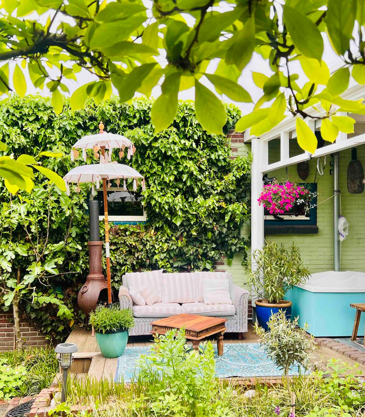 A comfortable seating area is surrounded by lush greenery, featuring a cozy couch and a decorative table. A colorful parasol provides shade, while vibrant flowers add a touch of color. The space includes a small jacuzzi, creating a tranquil outdoor retreat.