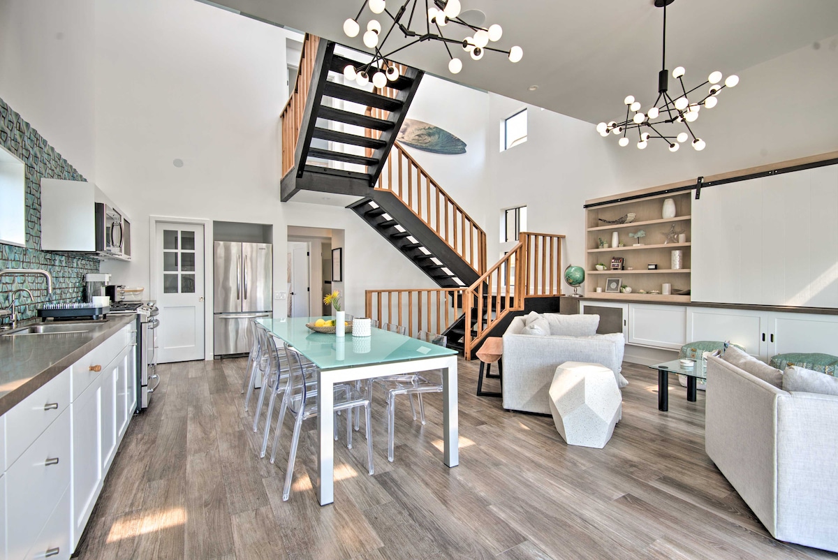 An open-concept living area features modern furnishings, including a stylish dining table surrounded by transparent chairs. A floating staircase ascends to a loft area, with natural light illuminating the space through large windows. The kitchen showcases contemporary appliances and sleek cabinetry.