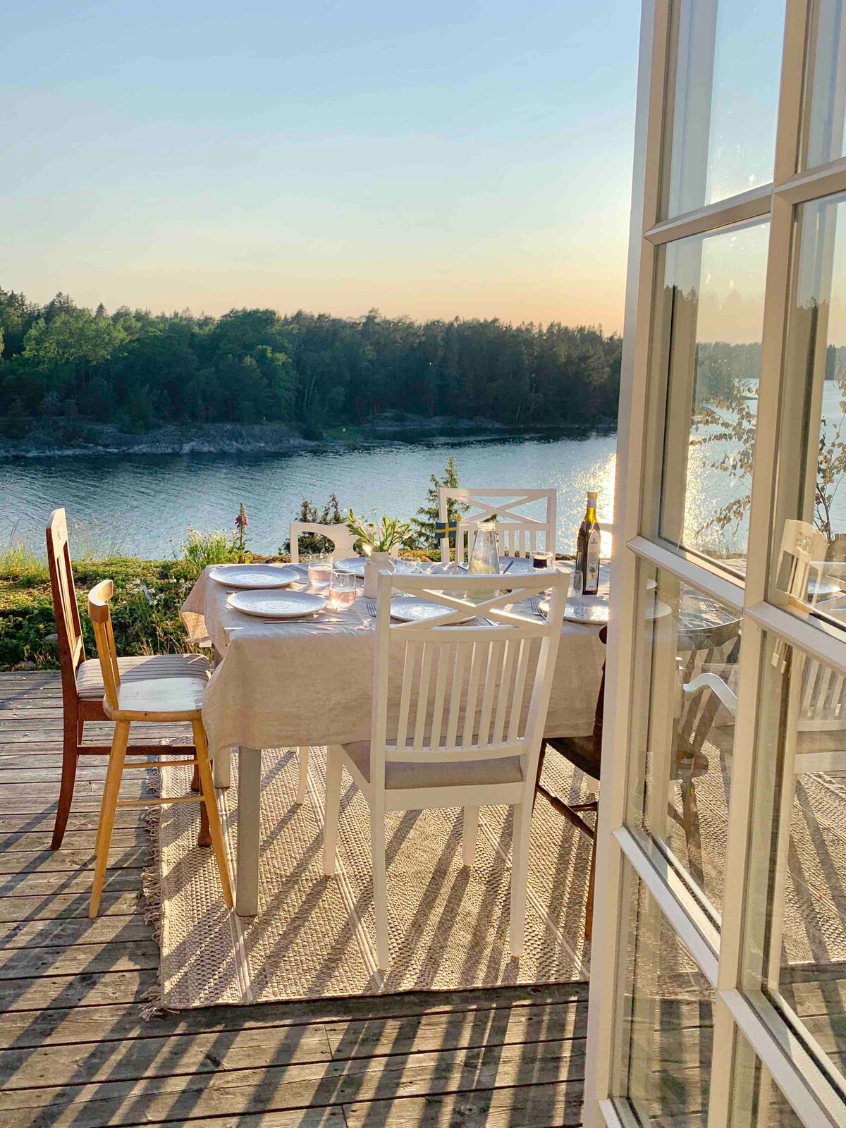 A dining area is set on a wooden terrace, overlooking serene water and lush greenery. The table is adorned with plates and glasses, while chairs surround it. Large glass doors provide a clear view of the tranquil scenery and sunset.