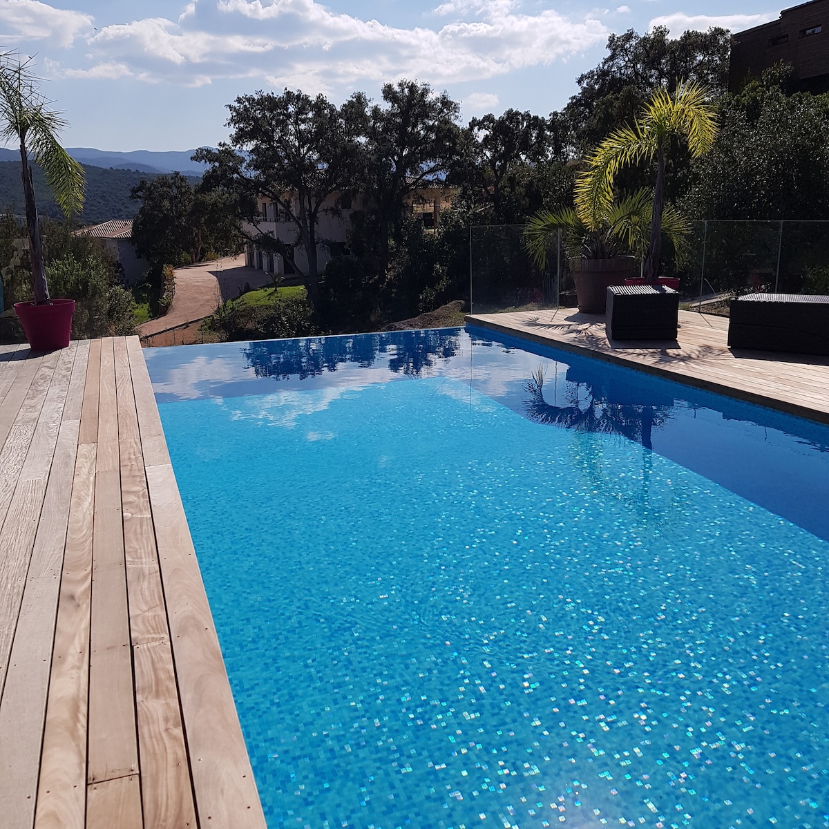 A heated pool is surrounded by a wooden deck, featuring a smooth surface that reflects the sky. Lush greenery is visible in the background, creating a serene atmosphere. Comfortable seating areas, consisting of several outdoor chairs, provide spaces for relaxation near the water.