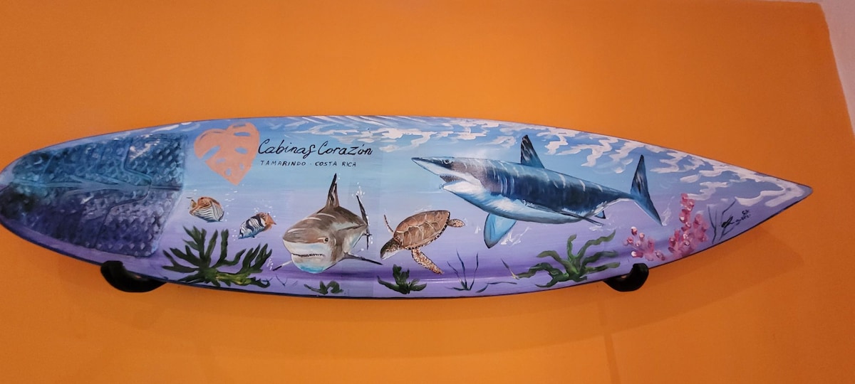 A surfboard is displayed against a vibrant orange wall, featuring hand-painted marine life, including a shark and a turtle, along with coral and underwater plants. The artwork reflects a coastal theme, enhancing the room's surfer-inspired atmosphere.